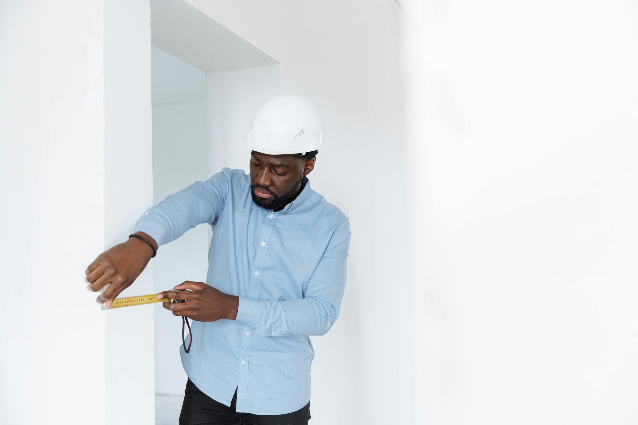 A man in a hard hat measures a wall, showcasing precision in a building interior.